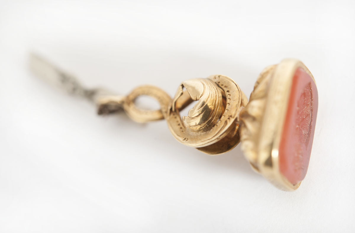 Sigill, guld, med sten av karneol. F.H. af Chapmans adelsvapen. Krönt, hjärtformad sköld med vinkelhake och tre månskäror å räfflad botten. Drivet och ciselerat guld med ring för urkedja. Tillhörande sigilavtryck.