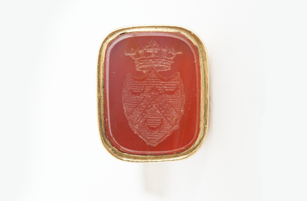 Sigill, guld, med sten av karneol. F.H. af Chapmans adelsvapen. Krönt, hjärtformad sköld med vinkelhake och tre månskäror å räfflad botten. Drivet och ciselerat guld med ring för urkedja. Tillhörande sigilavtryck.