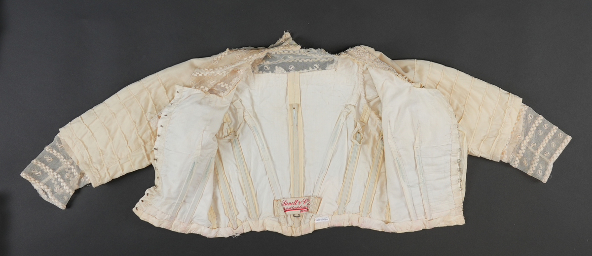 Del av drakt.  Overdel. Jakke.  Kvit med gulskjær. Jakka har tyllhals og tyllmansjettar. Tylldelen i halsområdet vert lukka til eine sida med åtte trykknapper. Blondekant rundt halsopninga og noko nedover på framstykket.  Opning midt framme. Lukking av fôr-delen med 13 hekter og hemper.  Lukking av ytterste stoff (framstykket) med sju trykknappar. I fôret er det sydd på 11 loddrette spilar (inni "lommer"), i midt i rygg, to på kvar side av bakstykket og to på kvar side av framstykket. 
Kvart erme er pynta med åtte sydde legg på ytste laget. Inni erma er det eit "laust" erme i fôrstoff (sydd fast i saum i ermholet). I enden av dette ermet er det påsydd blondestoff (same som rundt halsopninga) som stikk ut i frå det ytterste ermet.