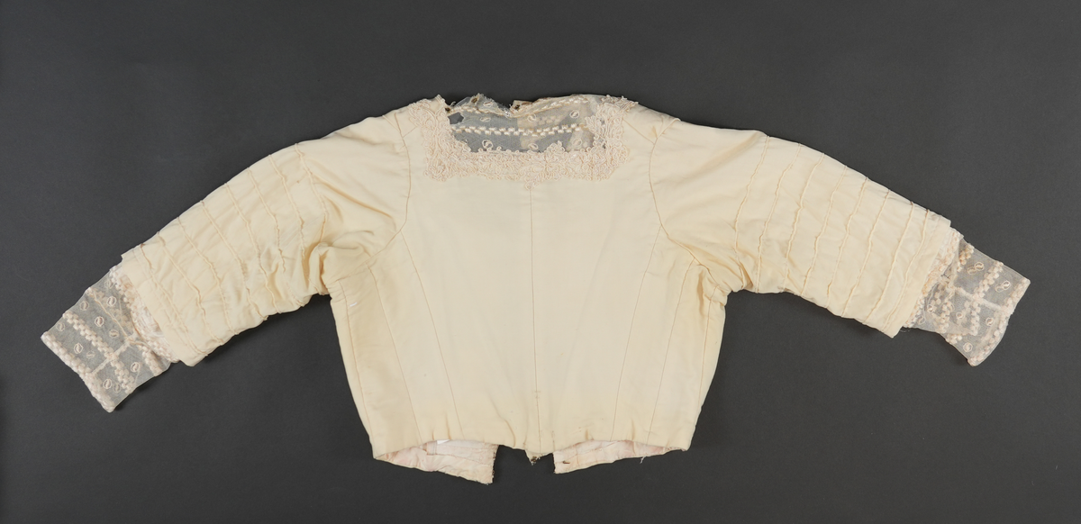 Del av drakt.  Overdel. Jakke.  Kvit med gulskjær. Jakka har tyllhals og tyllmansjettar. Tylldelen i halsområdet vert lukka til eine sida med åtte trykknapper. Blondekant rundt halsopninga og noko nedover på framstykket.  Opning midt framme. Lukking av fôr-delen med 13 hekter og hemper.  Lukking av ytterste stoff (framstykket) med sju trykknappar. I fôret er det sydd på 11 loddrette spilar (inni "lommer"), i midt i rygg, to på kvar side av bakstykket og to på kvar side av framstykket. 
Kvart erme er pynta med åtte sydde legg på ytste laget. Inni erma er det eit "laust" erme i fôrstoff (sydd fast i saum i ermholet). I enden av dette ermet er det påsydd blondestoff (same som rundt halsopninga) som stikk ut i frå det ytterste ermet.