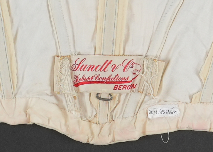 Del av drakt.  Overdel. Jakke.  Kvit med gulskjær. Jakka har tyllhals og tyllmansjettar. Tylldelen i halsområdet vert lukka til eine sida med åtte trykknapper. Blondekant rundt halsopninga og noko nedover på framstykket.  Opning midt framme. Lukking av fôr-delen med 13 hekter og hemper.  Lukking av ytterste stoff (framstykket) med sju trykknappar. I fôret er det sydd på 11 loddrette spilar (inni "lommer"), i midt i rygg, to på kvar side av bakstykket og to på kvar side av framstykket. 
Kvart erme er pynta med åtte sydde legg på ytste laget. Inni erma er det eit "laust" erme i fôrstoff (sydd fast i saum i ermholet). I enden av dette ermet er det påsydd blondestoff (same som rundt halsopninga) som stikk ut i frå det ytterste ermet.