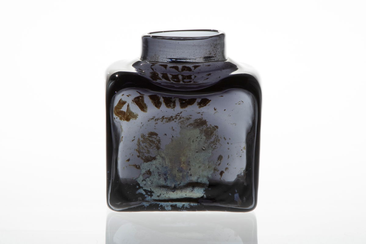Firkantet vase i mørkt glass. Vasen har smal, lav og rund åpning. Blåst i form. På to sider er det mønster i sølvfarget, iriserende ilegg.