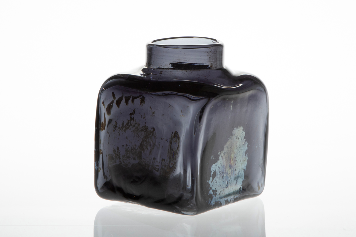 Firkantet vase i mørkt glass. Vasen har smal, lav og rund åpning. Blåst i form. På to sider er det mønster i sølvfarget, iriserende ilegg.