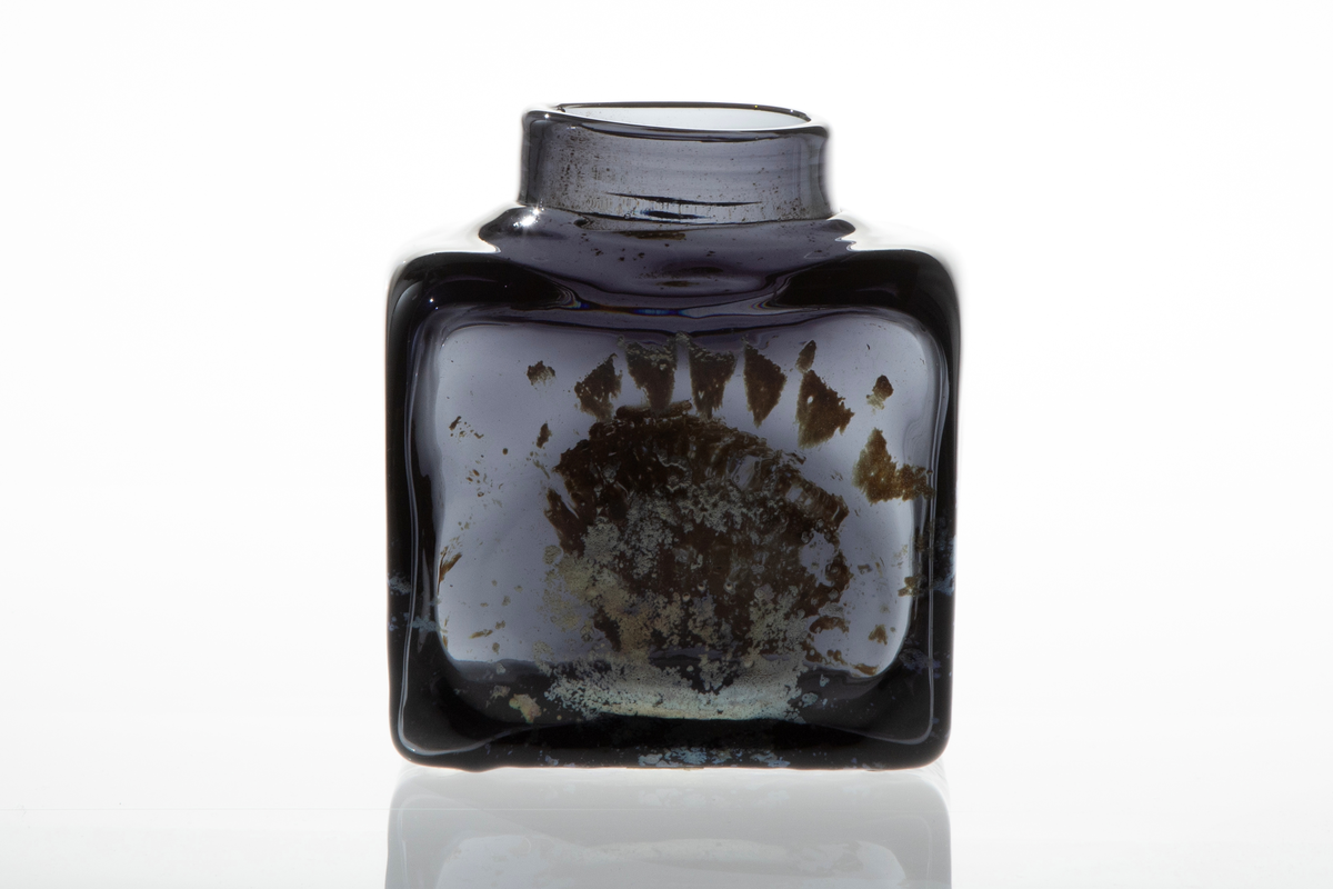 Firkantet vase i mørkt glass. Vasen har smal, lav og rund åpning. Blåst i form. På to sider er det mønster i sølvfarget, iriserende ilegg.