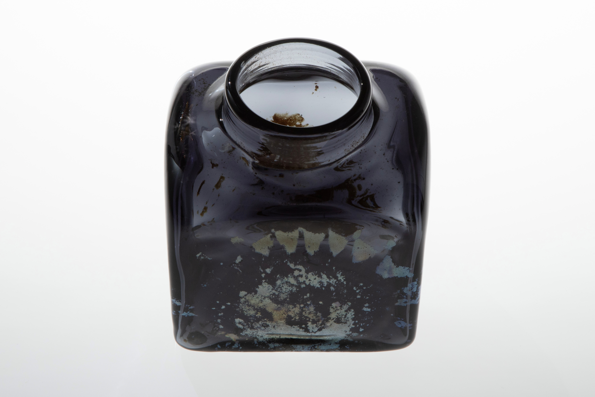 Firkantet vase i mørkt glass. Vasen har smal, lav og rund åpning. Blåst i form. På to sider er det mønster i sølvfarget, iriserende ilegg.