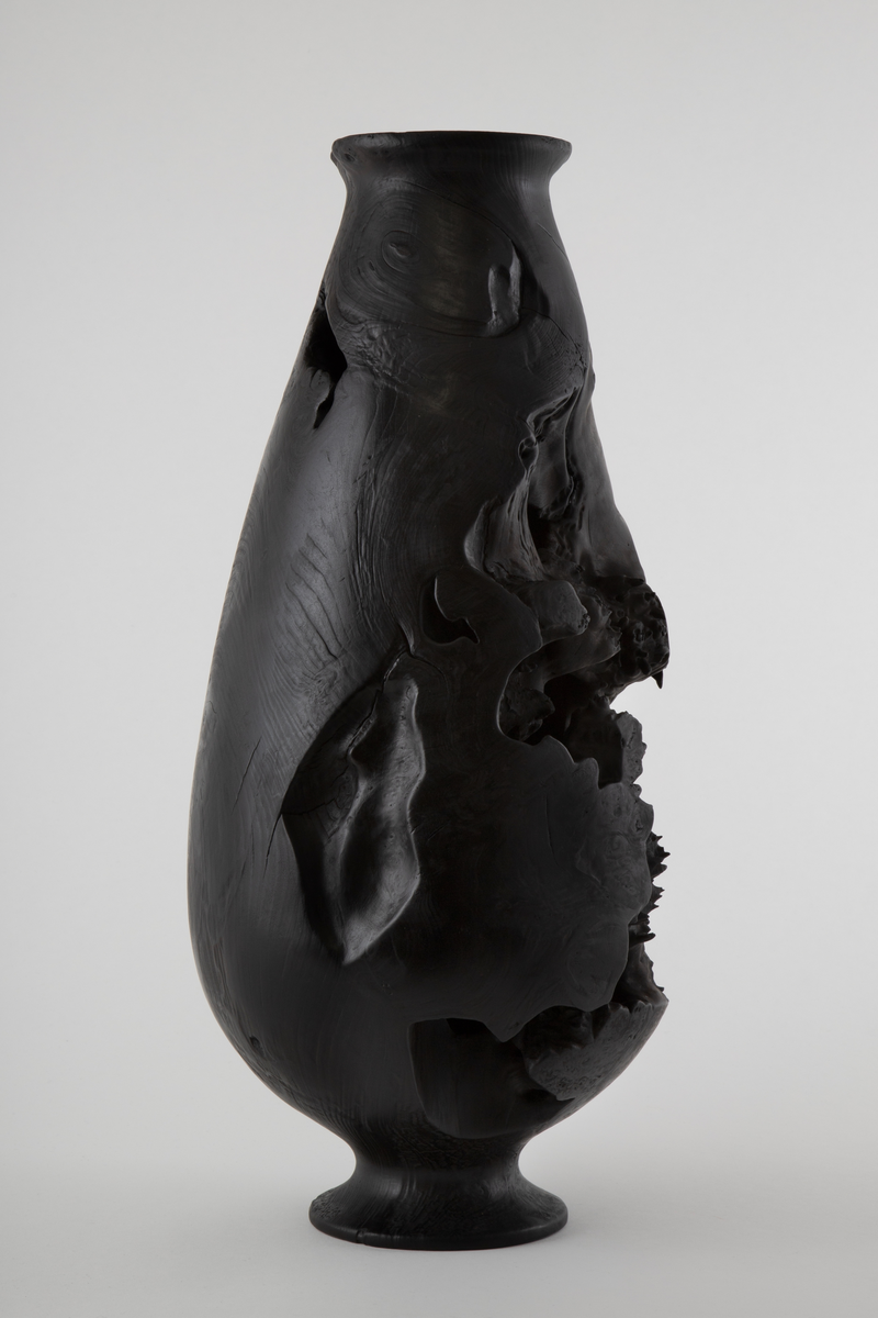 Vaseformet skulptur lagd av knoll fra hestekastanje. Høy og avlang form. Svartbrent med oljevoks.