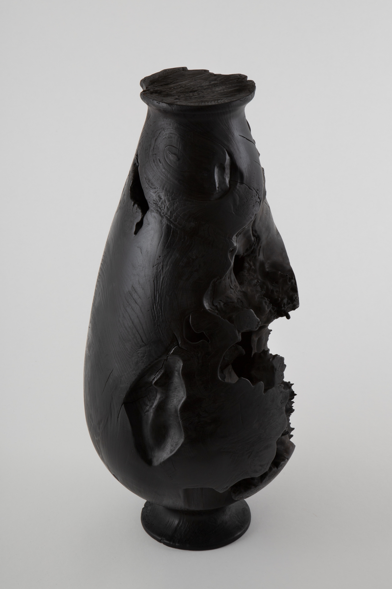 Vaseformet skulptur lagd av knoll fra hestekastanje. Høy og avlang form. Svartbrent med oljevoks.
