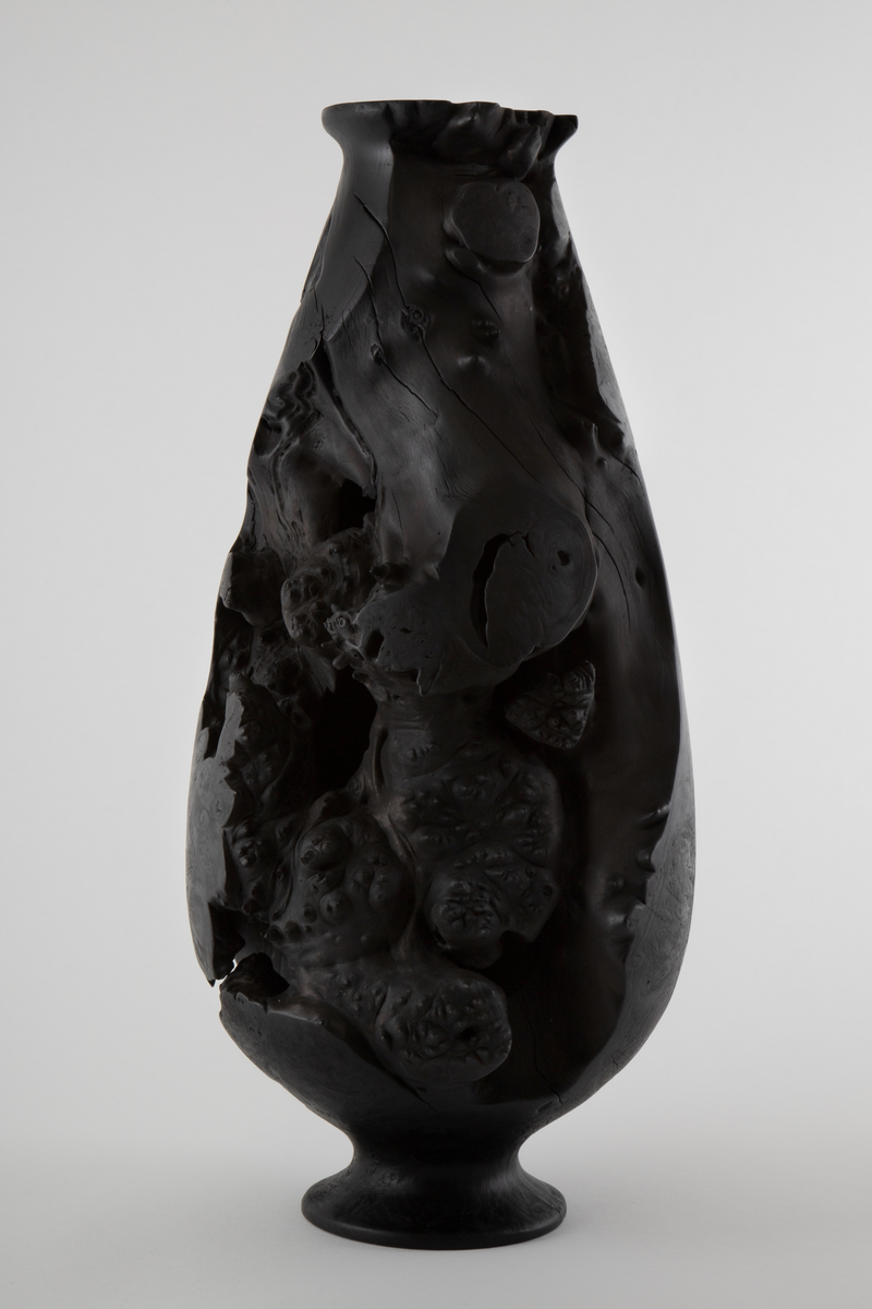 Vaseformet skulptur lagd av knoll fra hestekastanje. Høy og avlang form. Svartbrent med oljevoks.