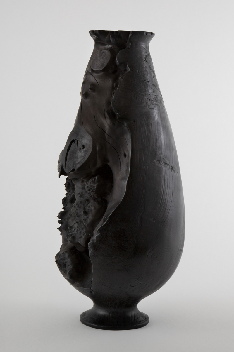 Vaseformet skulptur lagd av knoll fra hestekastanje. Høy og avlang form. Svartbrent med oljevoks.