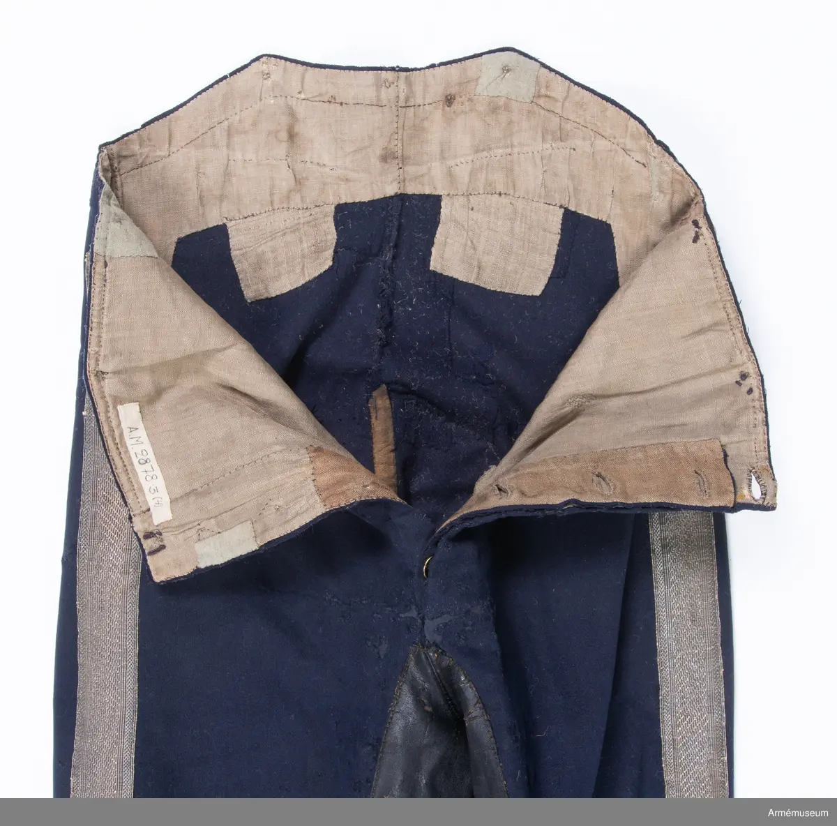 Mörkblå långbyxor med skinnskoning i baken och på benens insida, samt revärer av metallgalon i sidorna.

Byxorna är sydda av mörkblått kläde och har 4 cm breda revärer av silvergalon på benens utsidor. De är ursprungligen av en äldre modell med hel lucka fram, men har sytts om till gylf mitt fram, troligen i samband med att gylf fastställdes för långbyxor 1845. I varje sida syns spår efter luckan i form av ett smalt insnitt, samt ett igensytt knapphål.
Gylfen har dold knäppning med 4 enkla svartlackade knappar.
Byxorna är skinnskodda med brunt skinn i baken och på benens insidor, men det syns även rester efter en rad stygn ovanför knät på båda ben som eventuellt kan vara spår efter en äldre skinnskoning.
Byxorna är ofodrade, men har en infodring av oblekt bomullstyg i linningen upptill och en bit ner på framstycket. Infodringen är lappad på flera ställen.
På vänster byxben vid knät syns en stor lagad reva i tyget. Nertill vid fållen sitter i vänster byxben en sekundär hälla av svart resårband, samt en sekundär svart plastknapp. Dessa har troligen satts dit i samband med att byxorna varit utställda.
På insidan av höger bens fåll sitter två små knappar för fastsättning av hälla. En lapp eller lagning är dessutom iskarvad vid höger knä.
Byxorna har 3 knappar bak och 4 fram för fastsättning av hängslen. De två knapparna på höger sida fram är kullriga mässingsknappar, medan övriga är 4-hålsknappar av svartlackad metall.