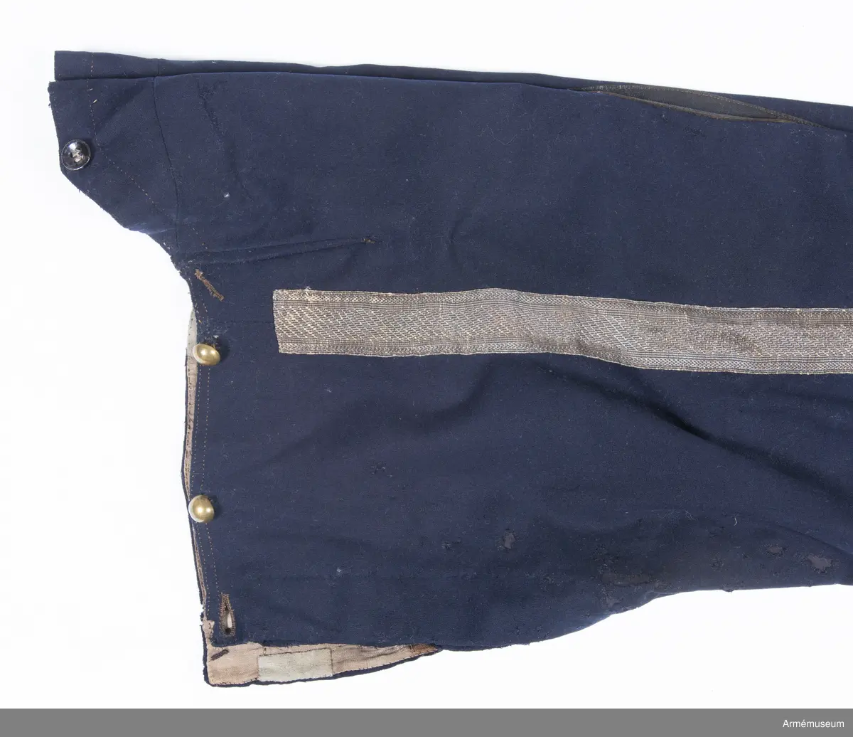 Mörkblå långbyxor med skinnskoning i baken och på benens insida, samt revärer av metallgalon i sidorna.

Byxorna är sydda av mörkblått kläde och har 4 cm breda revärer av silvergalon på benens utsidor. De är ursprungligen av en äldre modell med hel lucka fram, men har sytts om till gylf mitt fram, troligen i samband med att gylf fastställdes för långbyxor 1845. I varje sida syns spår efter luckan i form av ett smalt insnitt, samt ett igensytt knapphål.
Gylfen har dold knäppning med 4 enkla svartlackade knappar.
Byxorna är skinnskodda med brunt skinn i baken och på benens insidor, men det syns även rester efter en rad stygn ovanför knät på båda ben som eventuellt kan vara spår efter en äldre skinnskoning.
Byxorna är ofodrade, men har en infodring av oblekt bomullstyg i linningen upptill och en bit ner på framstycket. Infodringen är lappad på flera ställen.
På vänster byxben vid knät syns en stor lagad reva i tyget. Nertill vid fållen sitter i vänster byxben en sekundär hälla av svart resårband, samt en sekundär svart plastknapp. Dessa har troligen satts dit i samband med att byxorna varit utställda.
På insidan av höger bens fåll sitter två små knappar för fastsättning av hälla. En lapp eller lagning är dessutom iskarvad vid höger knä.
Byxorna har 3 knappar bak och 4 fram för fastsättning av hängslen. De två knapparna på höger sida fram är kullriga mässingsknappar, medan övriga är 4-hålsknappar av svartlackad metall.