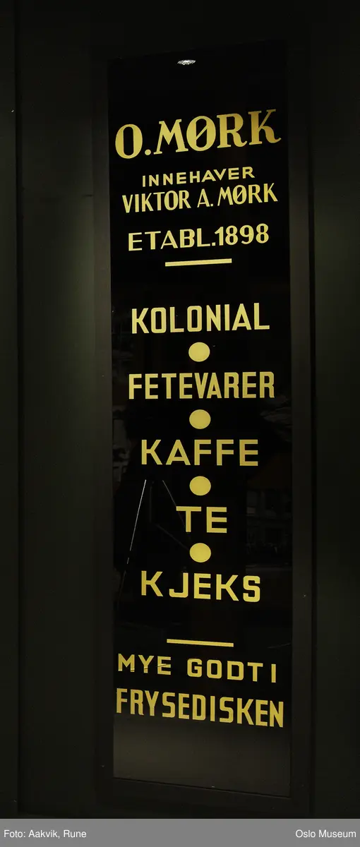 Butikkskilt i glass, sort med gylne bostaver. Sprukket, men konsolidert med plate bak.