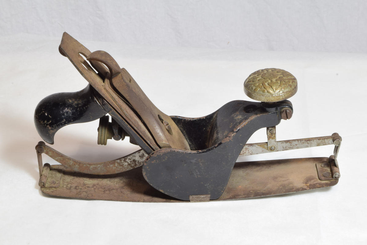 Justerbar buehøvel i metall. Håndtaket foran er dekorert med blomsterornamentikk. 
