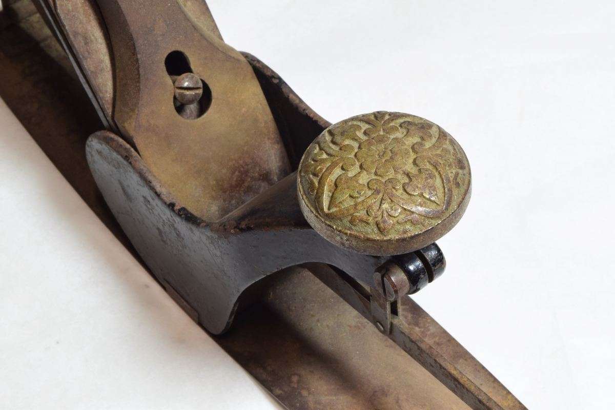 Justerbar buehøvel i metall. Håndtaket foran er dekorert med blomsterornamentikk. 
