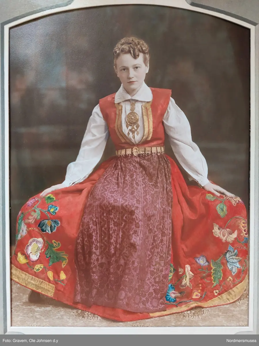 Brudedrakt med skjørt, liv, skjorte, forkle, stølebelte og offerdåse. 
Rødt bunadsliv av nordmørstype i glanset ullstoff med ufarget for. "Sølvkanter" langs åpningen på begge sider. 
Linskjorte der nedre del er av grovere tekstil. Øvre del har svært bred krage fullbrodert med hvitsøm og ermer med hvitsøm. 
Skjørt i rødt glanset ullstoff, fronten har innsydd et rødt ullstoff i en stripe som dekkes av forkleet. Langs nedre kant en maskinlaget silkebord. Omfattende appliklasjonsbroderier  i blomstermønster over store deler av nedre del  av skjørtet. Skjørtet er sydd sammen av flere deler av samme tekstil.
Offerdåse av vanlig modell av sølv med utklipt malteserkors hengende fra bunnen, hengende ruterformede sølvstykker fra hver side og bunnen av korset og fra selve offerdåsen på hver side av korset. Offerdåsen er på kjede som avslutter i to ruterformede figuerer med løkker til anheng på vher av ruterens sider. Inngravert geometrisk mønster.
Rødt stølebelte med støler av nordmørstype, mønster i form av tre talglys, i forgylt sølv med beltespenne av forgylt sølv. Fra hver av stølene henger to flate ringer.
Forkle med alternerende nedagående blomster og sikk-sakkmønster i silkestoff.