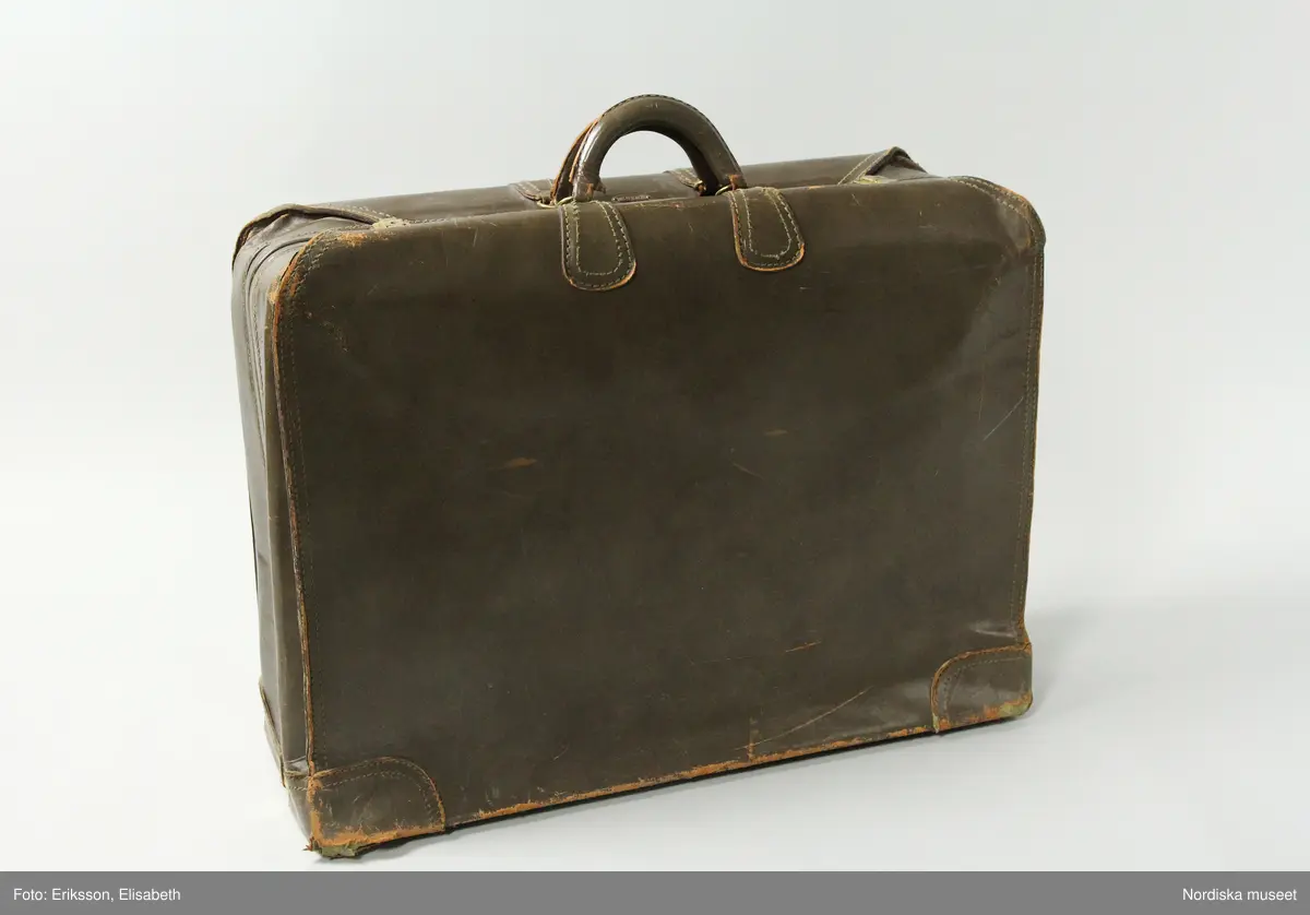 Resväska av gråbrunt läder, rektangulär, cirka 70 x 50 cm, med två styva handtag klädda med läder, fästa i ringar av gulmetall och förstärkningar av läder. Två knäppen av gulmetall stänger/låser väskan, nyckelhål finns (nyckel saknas). På ovansidan intill handtagen initialerna G. M. präglat i guldfärg samt en metallplakett med företagsnamnet SAKS FIFTH AVENUE. Undersidans fyra hörn förstärkta med läder, undertill fem kulfötter av gulmetall (den sjätte saknas). Väskan hopsydd med kraftiga, synliga maskinsömmar, bitvis med två parallella sömmar. Öppnas/delas på mitten i två lika stora hälfter, djup 10-12 cm vardera. Insidan klädd med tuskaftat, oblekt linnetyg, till vardera halvan en täckduk av bandkantat linnetyg, fäst i botten och mot ena sidan fäst med en spänntamp av linnetyg och spänne av gulmetall, mot andra sidan fäst med två vridbara knäppen av gulmetall. Ena täckduken försedd med ett stort fack med skinnskott blixtlås av gulmetall, löparen märkt SERVAL USA.
Anmärkning:
Utsidan repig och sliten med kraftigt slitna hörn, insidans linneinredning helt intakt. Nyckel samt en kulfot saknas.
/Marianne Larsson 2024-08-20