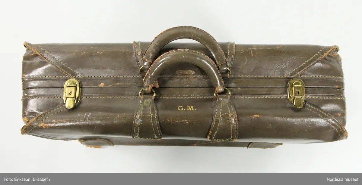 Resväska av gråbrunt läder, rektangulär, cirka 70 x 50 cm, med två styva handtag klädda med läder, fästa i ringar av gulmetall och förstärkningar av läder. Två knäppen av gulmetall stänger/låser väskan, nyckelhål finns (nyckel saknas). På ovansidan intill handtagen initialerna G. M. präglat i guldfärg samt en metallplakett med företagsnamnet SAKS FIFTH AVENUE. Undersidans fyra hörn förstärkta med läder, undertill fem kulfötter av gulmetall (den sjätte saknas). Väskan hopsydd med kraftiga, synliga maskinsömmar, bitvis med två parallella sömmar. Öppnas/delas på mitten i två lika stora hälfter, djup 10-12 cm vardera. Insidan klädd med tuskaftat, oblekt linnetyg, till vardera halvan en täckduk av bandkantat linnetyg, fäst i botten och mot ena sidan fäst med en spänntamp av linnetyg och spänne av gulmetall, mot andra sidan fäst med två vridbara knäppen av gulmetall. Ena täckduken försedd med ett stort fack med skinnskott blixtlås av gulmetall, löparen märkt SERVAL USA.
Anmärkning:
Utsidan repig och sliten med kraftigt slitna hörn, insidans linneinredning helt intakt. Nyckel samt en kulfot saknas.
/Marianne Larsson 2024-08-20