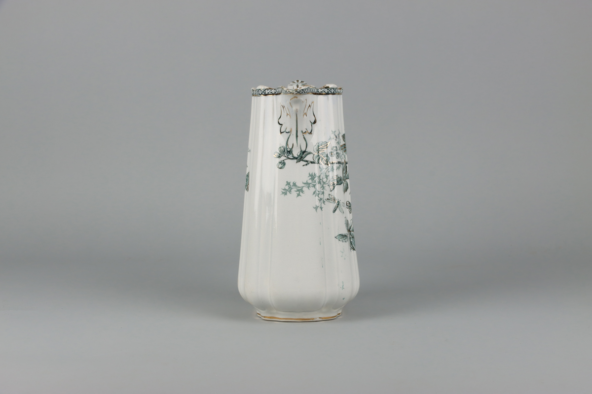 Kanna med pip och hänkel. Rektangulär form med något buktande och brutna rundade hörn. Upptill mot mynningen bård med kryssmönster. Under pip dekor med löv och på sidor dekor med fågel, växt och blommande kvistar. Dekor i grönt och guld på vitt grund. Nedtill låg profilerad fot. Under tryckt stämpel i grönt med arkitekturmotiv och servisnamn Weixiö Gustavsberg samt djupstämpel ES och siffran 2.



