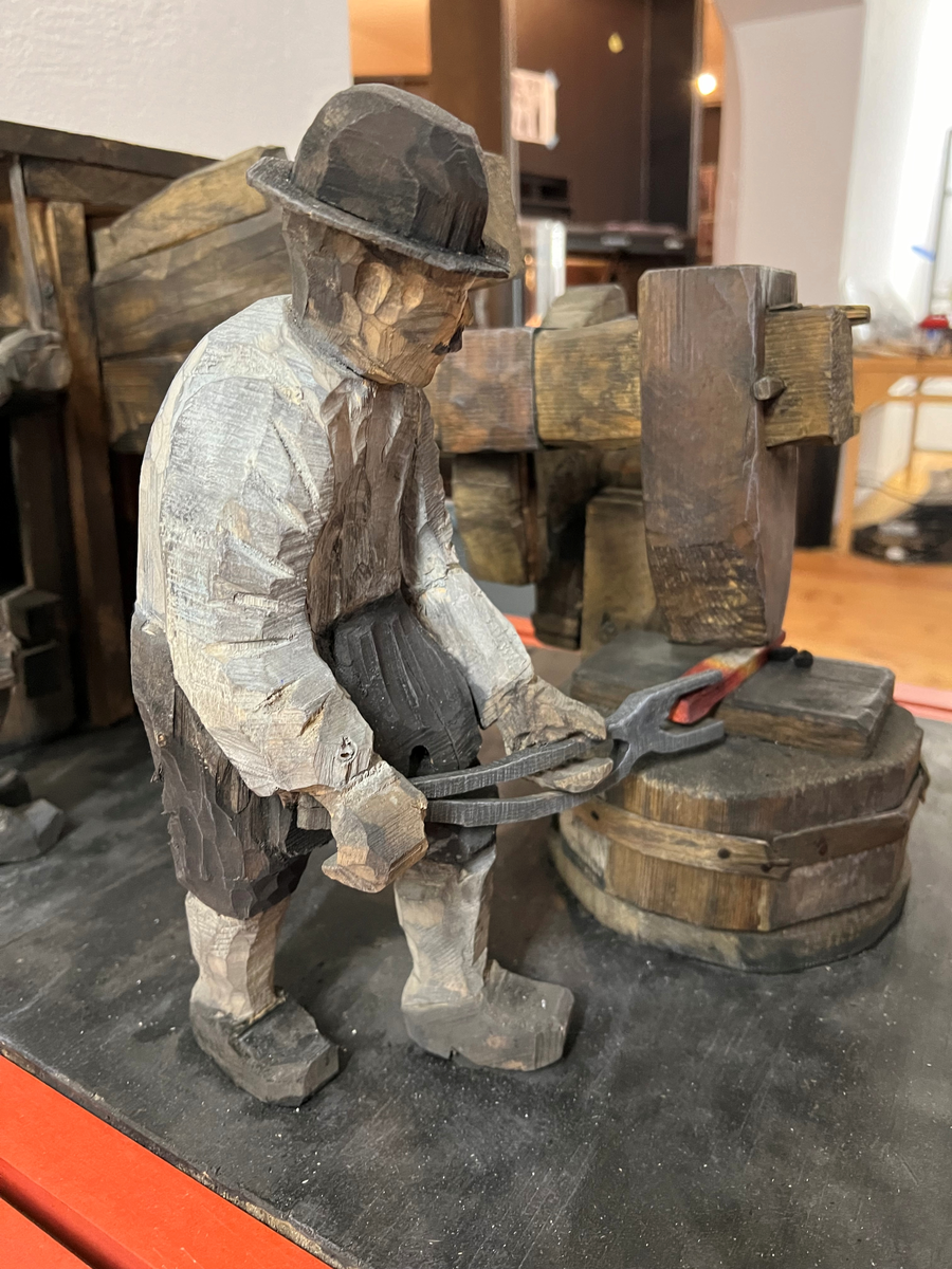 Skulptur av trä. "Stångjärnshammare", tillverkad av Hans Lustig.

Skulpturscen som framställer del av hammarsmedja med hammare och två män, en smed hållande järnstycket under hammaren, den andre sköter hammaren. Hela scenen på en stor platta.