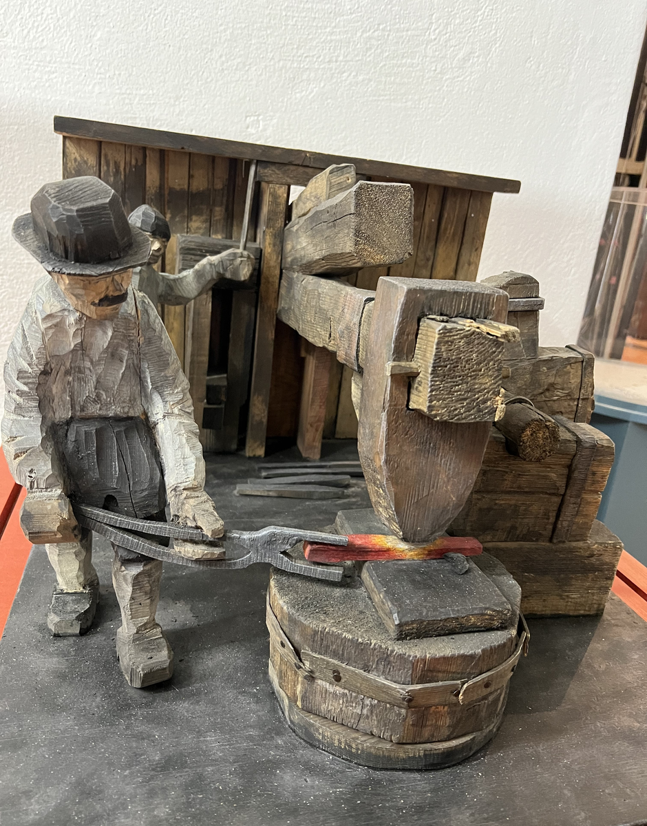 Skulptur av trä. "Stångjärnshammare", tillverkad av Hans Lustig.

Skulpturscen som framställer del av hammarsmedja med hammare och två män, en smed hållande järnstycket under hammaren, den andre sköter hammaren. Hela scenen på en stor platta.