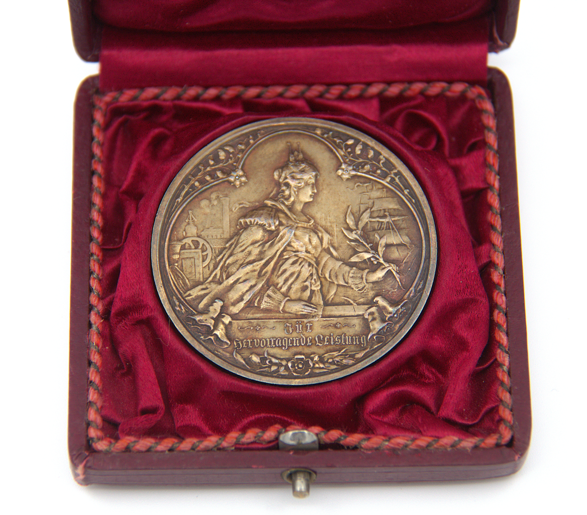 Medalj ur Ahlgrens medaljsamling. Tilldelades Ahlgrens Tekniska Fabrik i Lübeck 1895 (´för framstående prestation´). Guldfärgad. Asken är i burgundyröd trä och har insidan klädd med ett sidenliknande material samt omslag av läderimitation (papper). På locket finns en fastklistrad äldre lapp med handskriven information om plats och datering: "Lübeck 1895". Tidigare märkning: 2a.