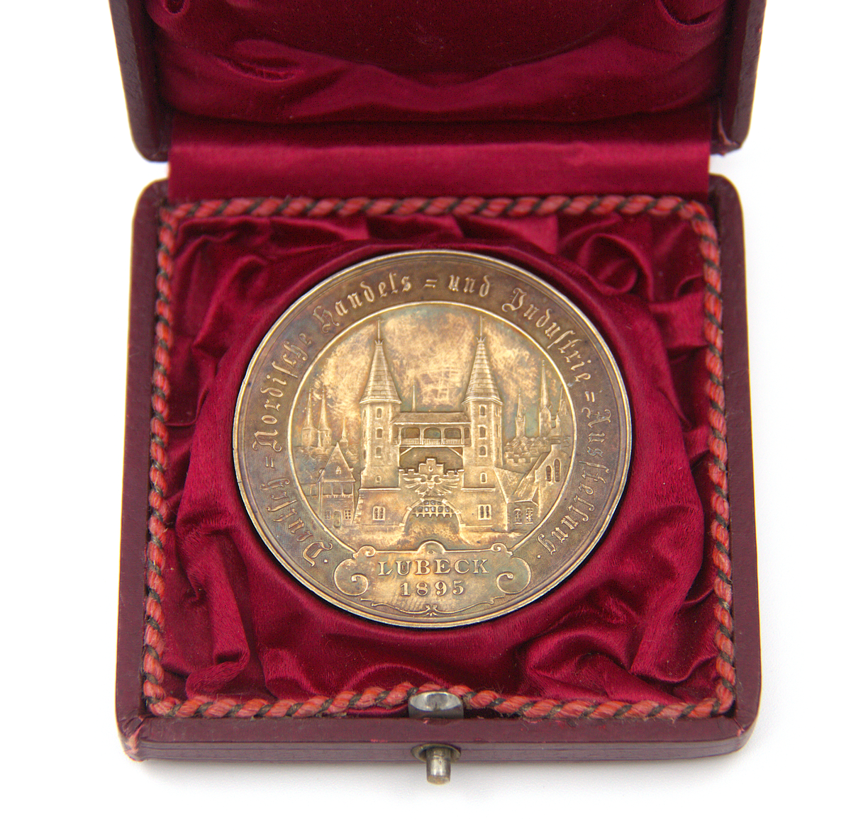 Medalj ur Ahlgrens medaljsamling. Tilldelades Ahlgrens Tekniska Fabrik i Lübeck 1895 (´för framstående prestation´). Guldfärgad. Asken är i burgundyröd trä och har insidan klädd med ett sidenliknande material samt omslag av läderimitation (papper). På locket finns en fastklistrad äldre lapp med handskriven information om plats och datering: "Lübeck 1895". Tidigare märkning: 2a.