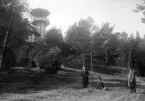 Två män och en kvinna i en park vid en högt belägen villa med torn. Okänd plats, ca. 1910-tal.