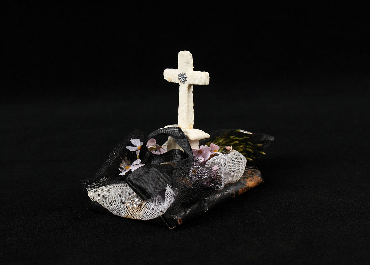 Begravningskaramell med svart tygrosett, pappersgräs, violetta blommor samt vitt, stående kors på svart platta. Korset förmodligen av sockermassa (morsell).
Pappersremsa med text undertill: "Säll är den som slutat har sin strid. Segerkronan fått och evig frid."