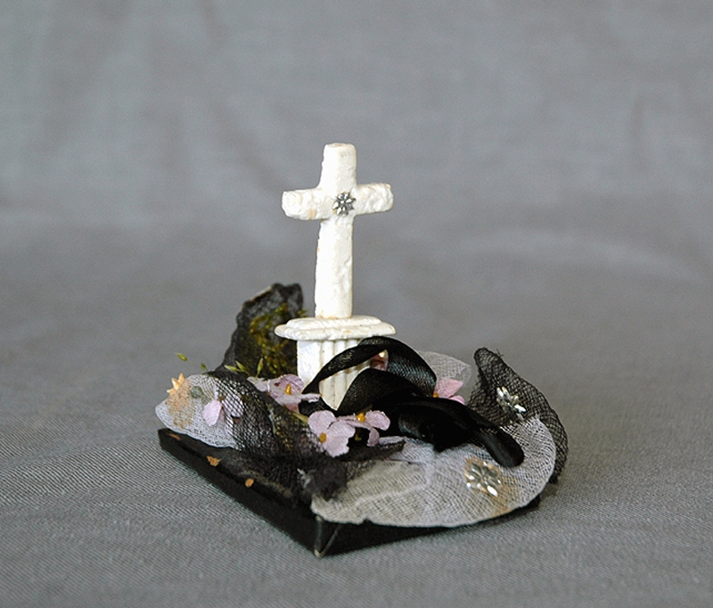 Begravningskaramell med svart tygrosett, pappersgräs, violetta blommor samt vitt, stående kors på svart platta. Korset förmodligen av sockermassa (morsell).
Pappersremsa med text undertill: "Säll är den som slutat har sin strid. Segerkronan fått och evig frid."