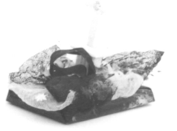 Begravningskaramell med svart tygrosett, pappersgräs, violetta blommor samt vitt, stående kors på svart platta. Korset förmodligen av sockermassa (morsell).
Pappersremsa med text undertill: "Säll är den som slutat har sin strid. Segerkronan fått och evig frid."