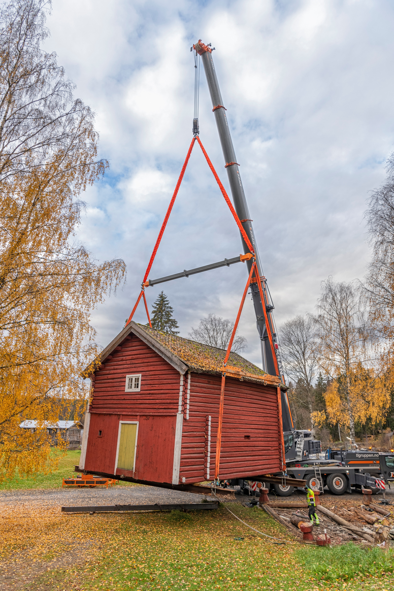 Flytting av stabburet på klokkergarden Fossum i Elverum, som fra tidlig på 1960-tallet har inngått i Norsk skogmuseums friluftsmusem (jfr. SJF-B.0020). Dette tunge løftet ble tatt som innledning til et restaureringsprosjekt som ble initiert fordi museets personale gjennom lengre tid hadde observert at svillramma og de ni cirka 70 centimeter høye stabbene under bygningen var råtne, og tilksynelatende i ferd med å kollapse. Ei 60 tonns mobilkran fra firmaet TL Kran ble plassert på vegen vest for stabburet. Deretter ble to kraftige H-bjelker av stål skjøvet inn under bygningen, på tvers. I disse bjelkenes ytterender ble det festet solide stropper, med stålstag som skulle forebygge at stålbjelkene forskjøv seg og at takutstikkene ble skadd. Dette fotografiet er tatt idet man så vidt hadde begynt å bevege bygningen i en halvsirkel over den nevnte vegen. 

Knut-Arild Nordli, som har spesialisert seg på antikvarisk restaureriring av laftekonstruksjoner, har påtatt seg å redde stabburet. Han vil støpe betongsåler med god bæreevne under bakkenivå, legge høvelig laftestein oppå som underlag for svillramma, som på grunn av råteskadene i stor utstrekning vil måtte bli en kopi av den gamle. Det tunge løftet ble gjort for at Knut-Arild skal kunne løse oppdraget sitt på en effektiv og trygg måte. Bygningen har ei grunnflate på 52 kvadratmeter og er to etasjer høy. Inkludert stålet i de nevnte H-bjelkene var løftet på cirka 32 tonn. Vi antar at sjølve bygningen veier drøyt 28 tonn. Den ble midlertidig flyttet til grassletta på den andre sida av vegen mot Klokkerfossbrua. Løftet gikk som det skulle. Det oppsto ingen nye skader. Når Nordli var ferdig med den nye og bedre fundamenterte underbygningen skulle stabburet – på samme måte – løftes tilbake til sin opprinnelige plass i Klokkergardstunet.