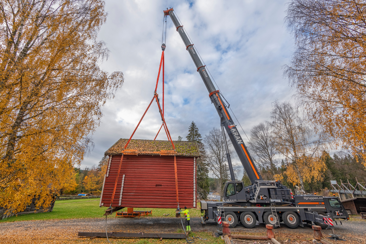 Flytting av stabburet på klokkergarden Fossum i Elverum, som fra tidlig på 1960-tallet har inngått i Norsk skogmuseums friluftsmusem (jfr. SJF-B.0020). Dette tunge løftet ble tatt som innledning til et restaureringsprosjekt som ble initiert fordi museets personale gjennom lengre tid hadde observert at svillramma og de ni cirka 70 centimeter høye stabbene under bygningen var råtne, og tilksynelatende i ferd med å kollapse. Ei 60 tonns mobilkran fra firmaet TL Kran ble plassert på vegen vest for stabburet. Deretter ble to kraftige H-bjelker av stål skjøvet inn under bygningen, på tvers. I disse bjelkenes ytterender ble det festet solide stropper, med stålstag som skulle forebygge at stålbjelkene forskjøv seg og at takutstikkene ble skadd. Dette fotografiet er tatt idet man så vidt hadde begynt å bevege bygningen i en halvsirkel over den nevnte vegen. Maskinen har en teleskoparm med en rekkevidde på opptil 60 meter. På slike distanser kan den bare løfte 6-700 kilo, men her, med objektet så nært kranbilen, var løftekapasiteten mye større.

Knut-Arild Nordli, som har spesialisert seg på antikvarisk restaureriring av laftekonstruksjoner, har påtatt seg å redde stabburet. Han vil støpe betongsåler med god bæreevne under bakkenivå, legge høvelig laftestein oppå som underlag for svillramma, som på grunn av råteskadene i stor utstrekning vil måtte bli en kopi av den gamle. Det tunge løftet ble gjort for at Knut-Arild skal kunne løse oppdraget sitt på en effektiv og trygg måte. Bygningen har ei grunnflate på 52 kvadratmeter og er to etasjer høy. Inkludert stålet i de nevnte H-bjelkene var løftet på cirka 32 tonn. Vi antar at sjølve bygningen veier drøyt 28 tonn. Den ble midlertidig flyttet til grassletta på den andre sida av vegen mot Klokkerfossbrua. Løftet gikk som det skulle. Det oppsto ingen nye skader. Når Nordli var ferdig med den nye og bedre fundamenterte underbygningen skulle stabburet – på samme måte – løftes tilbake til sin opprinnelige plass i Klokkergardstunet.