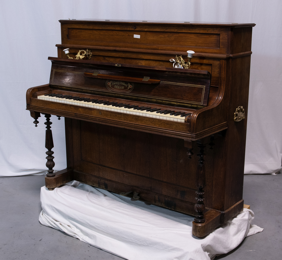 Piano med stående pianokasse i finert jakaranda med pianolokk, lite topplokk og to dreide, fremre konsollben. På sidene er to bærehåndtak i messing. Over pianolokket er to lysholdere i messing og porselen. To pedaler.