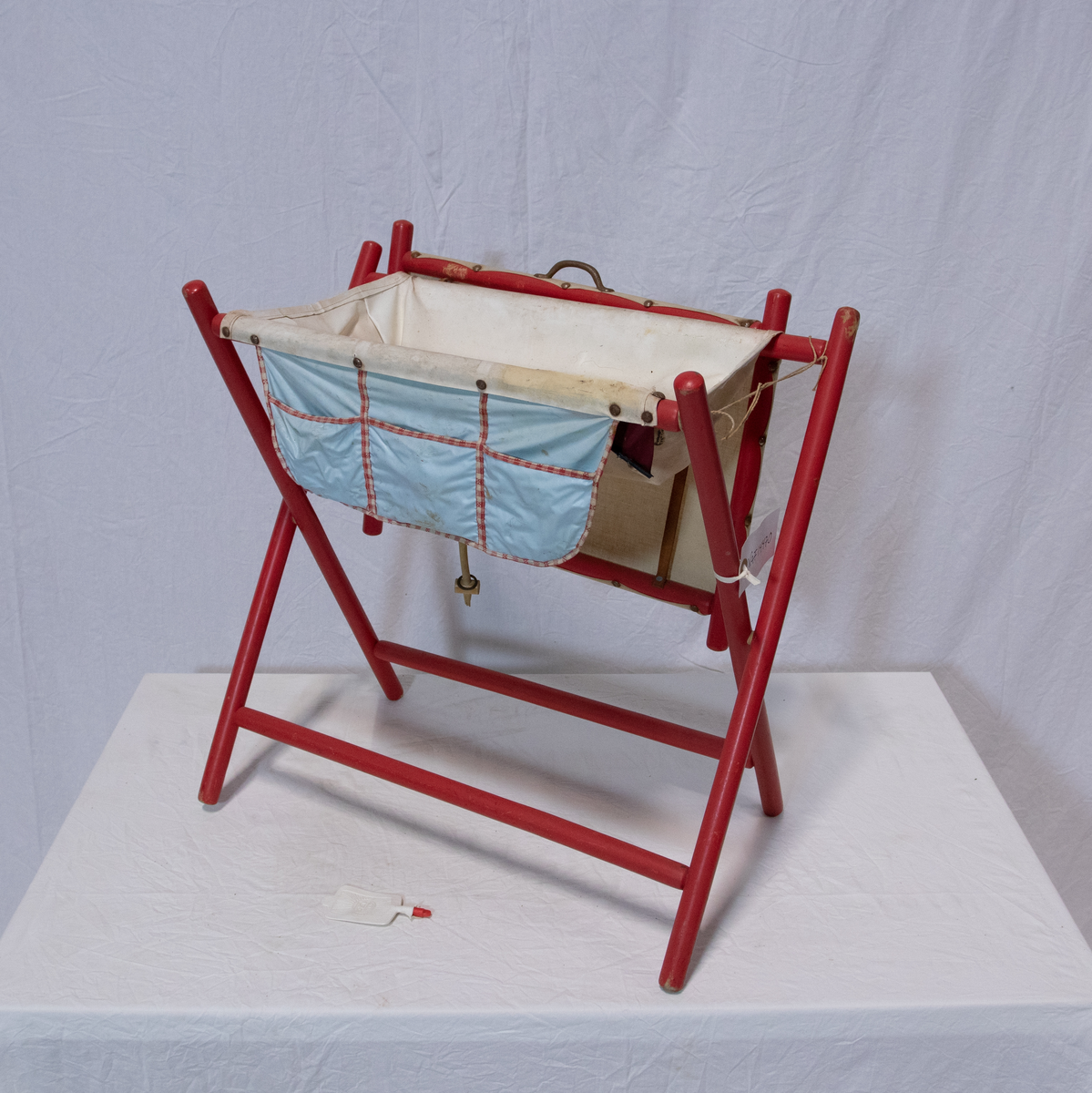 Stellebord til dukke. Rødmalt trekonstruksjon av runde pinner, tappet sammen i kryss i bena. Kan slås sammen. Badekar naglet fast i trerammen, laget av plast/vinyl. Badekaret har propp med slange i bunnen. Stellematte kan legges oppå badekaret, denne er laget i bomull. Stellematte stives av ved to lærstropper under, disse er spikret fast. Påfestet bærhåndtak i toppen av metall, skrudd med to skruer. På ene siden ett stykke med lysblå plast med tre lommer, hele stykket og lommene er kantet med rødrutete stoff.