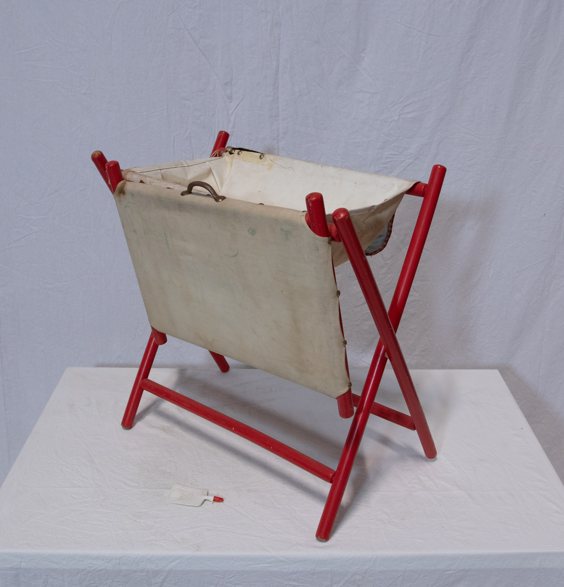 Stellebord til dukke. Rødmalt trekonstruksjon av runde pinner, tappet sammen i kryss i bena. Kan slås sammen. Badekar naglet fast i trerammen, laget av plast/vinyl. Badekaret har propp med slange i bunnen. Stellematte kan legges oppå badekaret, denne er laget i bomull. Stellematte stives av ved to lærstropper under, disse er spikret fast. Påfestet bærhåndtak i toppen av metall, skrudd med to skruer. På ene siden ett stykke med lysblå plast med tre lommer, hele stykket og lommene er kantet med rødrutete stoff.