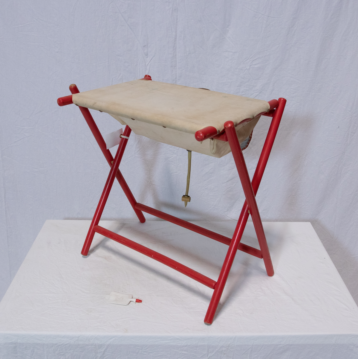 Stellebord til dukke. Rødmalt trekonstruksjon av runde pinner, tappet sammen i kryss i bena. Kan slås sammen. Badekar naglet fast i trerammen, laget av plast/vinyl. Badekaret har propp med slange i bunnen. Stellematte kan legges oppå badekaret, denne er laget i bomull. Stellematte stives av ved to lærstropper under, disse er spikret fast. Påfestet bærhåndtak i toppen av metall, skrudd med to skruer. På ene siden ett stykke med lysblå plast med tre lommer, hele stykket og lommene er kantet med rødrutete stoff.