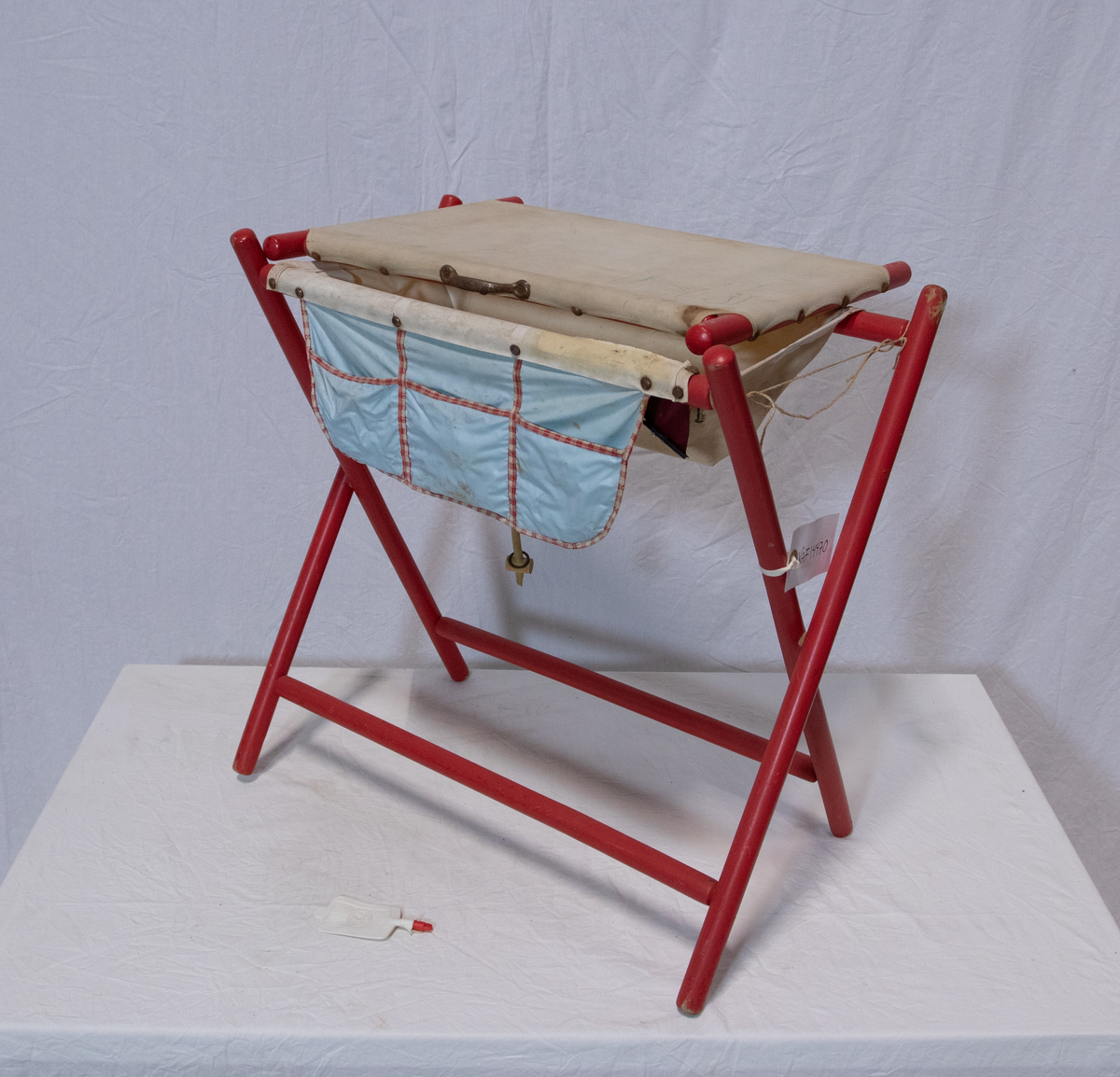 Stellebord til dukke. Rødmalt trekonstruksjon av runde pinner, tappet sammen i kryss i bena. Kan slås sammen. Badekar naglet fast i trerammen, laget av plast/vinyl. Badekaret har propp med slange i bunnen. Stellematte kan legges oppå badekaret, denne er laget i bomull. Stellematte stives av ved to lærstropper under, disse er spikret fast. Påfestet bærhåndtak i toppen av metall, skrudd med to skruer. På ene siden ett stykke med lysblå plast med tre lommer, hele stykket og lommene er kantet med rødrutete stoff.