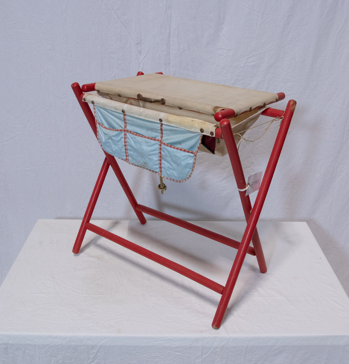 Stellebord til dukke. Rødmalt trekonstruksjon av runde pinner, tappet sammen i kryss i bena. Kan slås sammen. Badekar naglet fast i trerammen, laget av plast/vinyl. Badekaret har propp med slange i bunnen. Stellematte kan legges oppå badekaret, denne er laget i bomull. Stellematte stives av ved to lærstropper under, disse er spikret fast. Påfestet bærhåndtak i toppen av metall, skrudd med to skruer. På ene siden ett stykke med lysblå plast med tre lommer, hele stykket og lommene er kantet med rødrutete stoff.