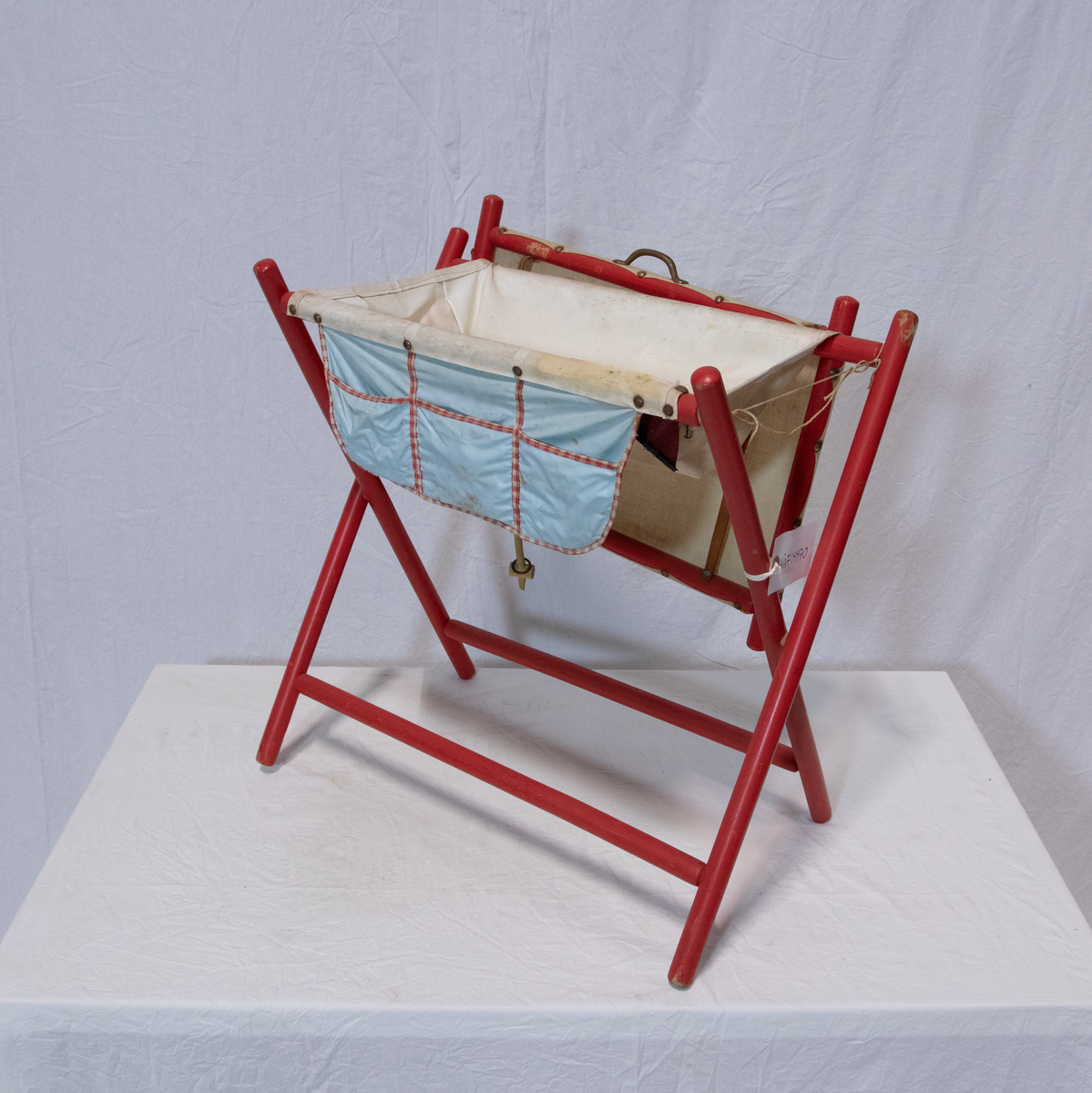 Stellebord til dukke. Rødmalt trekonstruksjon av runde pinner, tappet sammen i kryss i bena. Kan slås sammen. Badekar naglet fast i trerammen, laget av plast/vinyl. Badekaret har propp med slange i bunnen. Stellematte kan legges oppå badekaret, denne er laget i bomull. Stellematte stives av ved to lærstropper under, disse er spikret fast. Påfestet bærhåndtak i toppen av metall, skrudd med to skruer. På ene siden ett stykke med lysblå plast med tre lommer, hele stykket og lommene er kantet med rødrutete stoff.