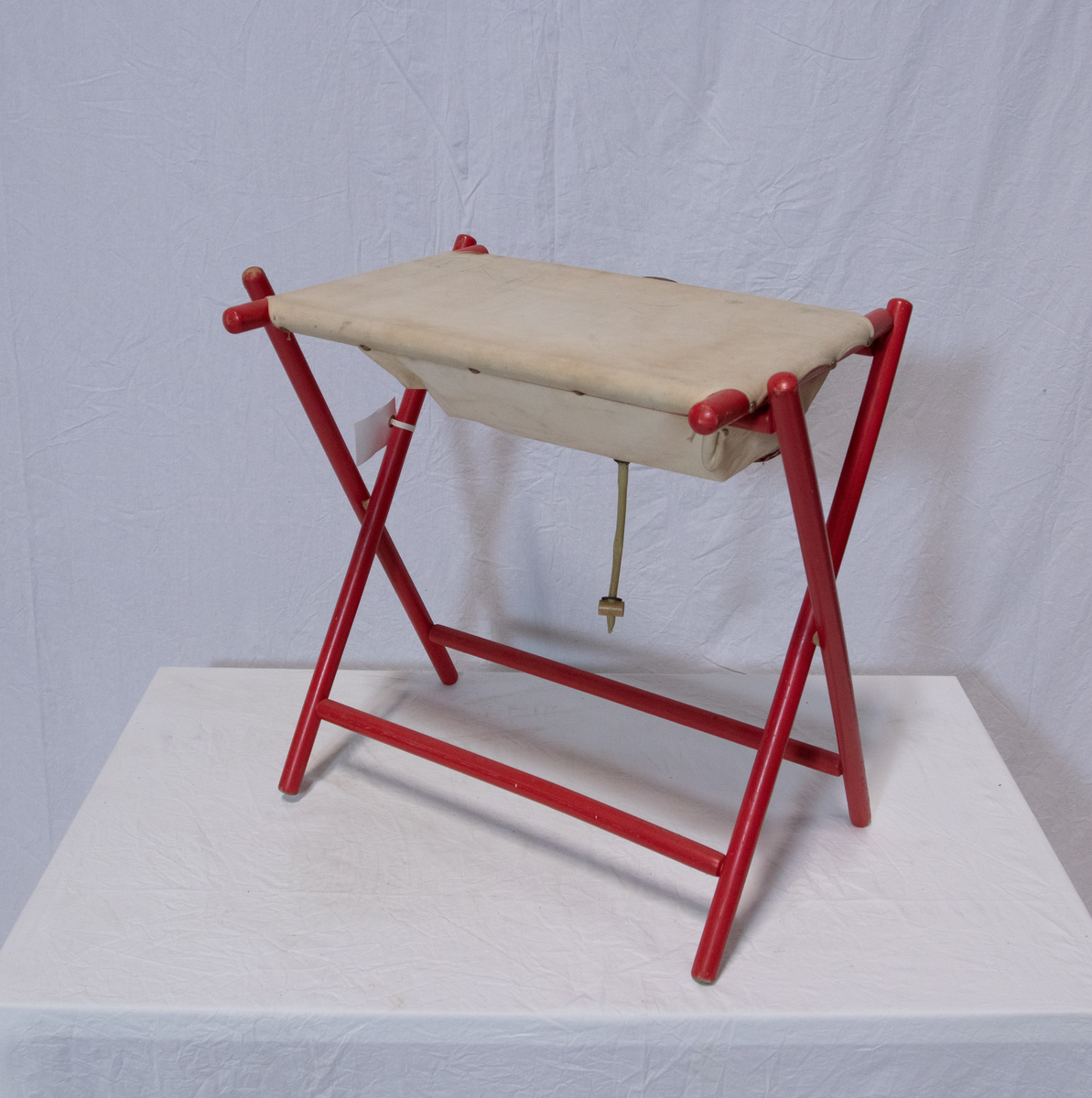 Stellebord til dukke. Rødmalt trekonstruksjon av runde pinner, tappet sammen i kryss i bena. Kan slås sammen. Badekar naglet fast i trerammen, laget av plast/vinyl. Badekaret har propp med slange i bunnen. Stellematte kan legges oppå badekaret, denne er laget i bomull. Stellematte stives av ved to lærstropper under, disse er spikret fast. Påfestet bærhåndtak i toppen av metall, skrudd med to skruer. På ene siden ett stykke med lysblå plast med tre lommer, hele stykket og lommene er kantet med rødrutete stoff.