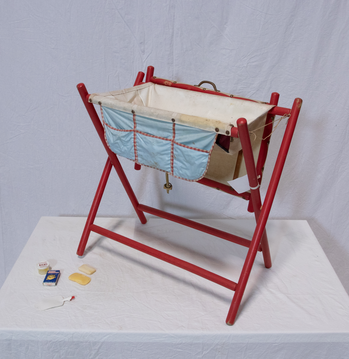 Stellebord til dukke. Rødmalt trekonstruksjon av runde pinner, tappet sammen i kryss i bena. Kan slås sammen. Badekar naglet fast i trerammen, laget av plast/vinyl. Badekaret har propp med slange i bunnen. Stellematte kan legges oppå badekaret, denne er laget i bomull. Stellematte stives av ved to lærstropper under, disse er spikret fast. Påfestet bærhåndtak i toppen av metall, skrudd med to skruer. På ene siden ett stykke med lysblå plast med tre lommer, hele stykket og lommene er kantet med rødrutete stoff.