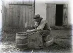 Jakob Söderlund slår bänden på laggkärlet. Det kan vara en insaltningstunna han arbetar med. I bakgrunden syns hans lilla ladugård, men den har ett tillbygge i trä t v som inte finns idag.