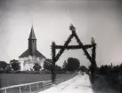 Till brudparet Elin och Anton Olsson Anderse förärades denna port framför Lau kyrka till sitt bröllop denna dag. Det syns att det var en blåsig dag! Folk är på väg till kyrkan för att vara med om vigseln.

Här ser man hur kyrkans gavel inte är färdigbyggd ända upp i sten, utan ifylld med provisoriska bräder, vilka måste ha förnyats åtskilliga gånger sedan kyrkan tillkomst på 1220-talet. Kanske blev inte heller taket färdigt, det är för flackt. 1958-60 byggde man upp det som fattades av gaveln med hålsten och putsade med cementputs. Tornet/takryttaren kom till på 1700-talet och byggdes märkligt nog lite utanför väggen. 1960 gjorde man en ny lite nättare takryttare.

Åkern framför kyrkan heter Stavgardsakarn. Stavgard är ett vanligt namn på järnålderns helgedomar, vilket visar att denna plats använts mycket länge för socknens behov.