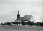 Masse har tagit denna bild stående i rågåkern söder om kyrkan. Han har medvetet valt motivet så att skolan inte finns med, dess östra hörn måste finnas alldeles utanför bildens kant t v. Handelsföreningens affär i vägskälet t h är ännu inte byggd. Kyrkan framträder monumentalt. Uppe på taket sitter ett gäng nyfikna ungdomar (?), hur har de kommit upp dit?