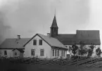 Masse har fotograferat skolan från söder med kyrkan i bakgrunden. Träden vid kyrkogårdsmuren är ännu små. Skolan är här 21 år gammal. Taket är täckt med falsad plåt i små ark, ett ovanligt takmaterial på landsbygden. Närmast ligger storskolan med tre fönster på långsidan och bakom småskolan med två fönster. T v är lärarbostäderna.