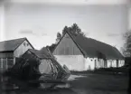 Här ser man att gårdstomten ligger lågt, höstregnen har mättat marken så vattnet flyter ovanpå. Den gamla gårdsplatsen öster om vägen låg lite högre.     Man ser att ladugården troligen hade två fähusdelar med en genomkörning emellan. Längst bort var det lada. Taket är täckt med halm och har vindskivor med vingliknande avslutningar. Boden är nog en vedbod, mot vars bakvägg det står en väldigt hop med stör till bandtunar.