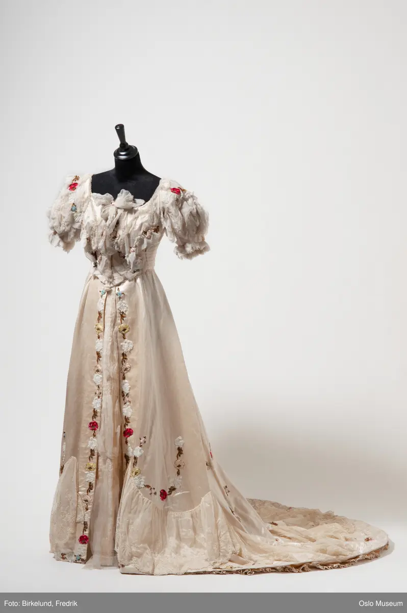 Overdel og skjørt. 
Kremfarget gallakjole i silkeduchesse. Applikert med maskinbroderte blomster, blader og perlefiguerer. innfelt chiffon over silkestoff stripet i moire og innvevet blomstermønster. Skjørtet er foret med organza. Overdel foret med tynn bomullslerret. Korte puffermer med chiffon og legg.