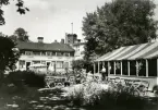 Café Centralparken i kvarteret Köpmannen, Hudiksvall, 1937. Huset byggt 1839. Till vänster syns en byggnad med många små fönster, vissa är öppna. Till höger syns två kvinnor framför en byggnad/paviljong med randig markis och blomsteramplar. Trädgårdsmöbler och växter på gården.