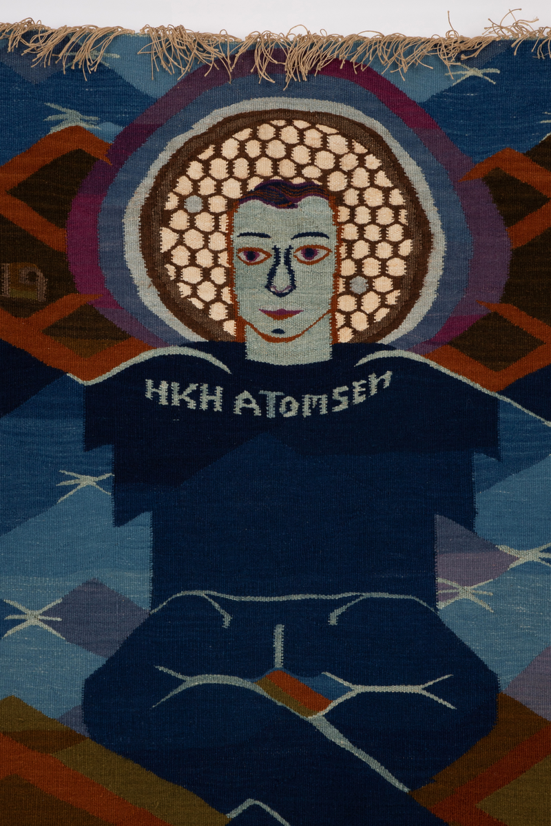 Billedvev med tre menneskefigurer. En blå figur sitter i lotusstilling med armene rettet utover. Han har en glorie rundt hodet. På brystet har han påskriften HKH Atomsen. Han er flankert av en hvitfarget naken mann og en kvinne på hver sin side. Kvinnen holder høyre hånd over brystet og med sin venstre hånd holder hun i hånden til en liggende person. Denne er blåfarget med røde omriss. Veven har også en del mønster.
