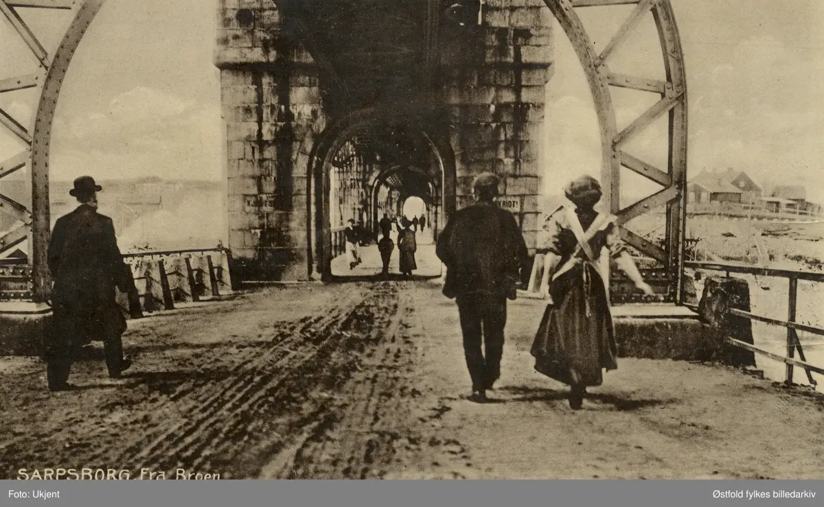 Sarpsbrua - under bruhvelvingene, Sarpsborg ca. 1900.
Fra Sarpsborgsiden, folk gående inn på brua - sett bakfra. Poskort.